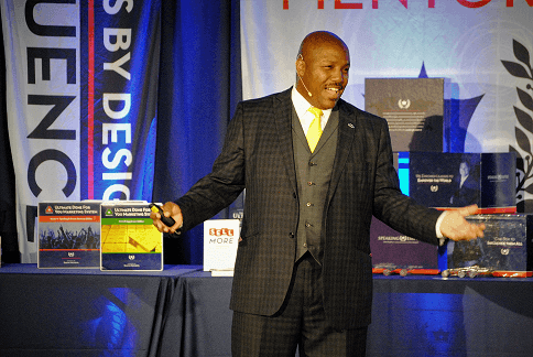 Man in a suit speaking on stage with a backdrop of banners and books on display tables.