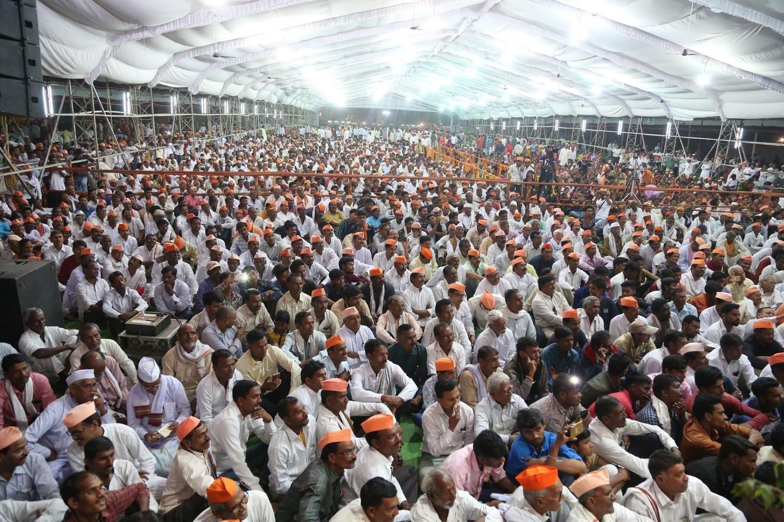 Large gathering of people seated under a tent, many wearing orange hats, at an event.